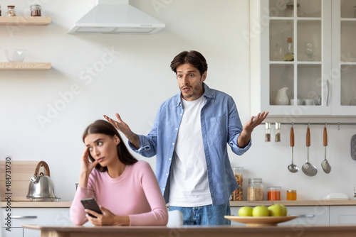 Unhappy young european lady with smartphone ignores offended angry screaming man at kitchen