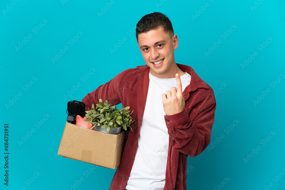 Young caucasian man making a move while picking up a box full of things isolated on blue background doing coming gesture Young caucasian man making a move while picking up a box full of things isolated on blue background doing coming gesture