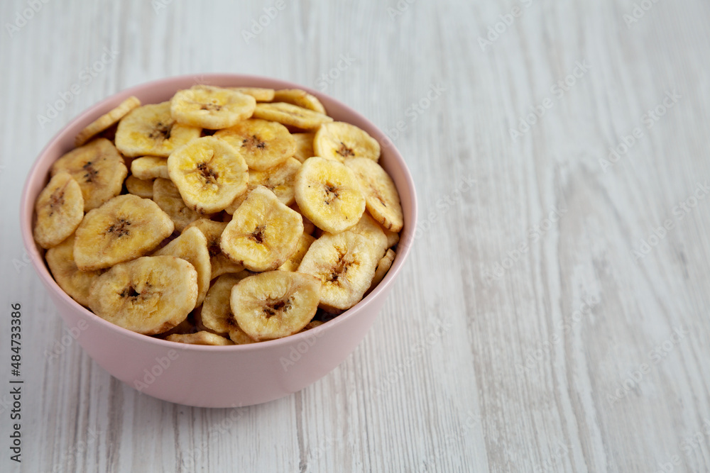 Homemade Banana Chips in a Pink Bowl on a white wooden background, side view. Space for text. Homemade Banana Chips in a Pink Bowl on a white wooden background, side view. Space for text.