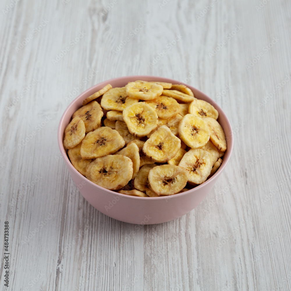 Homemade Banana Chips in a Pink Bowl on a white wooden background, side view. Homemade Banana Chips in a Pink Bowl on a white wooden background, side view.