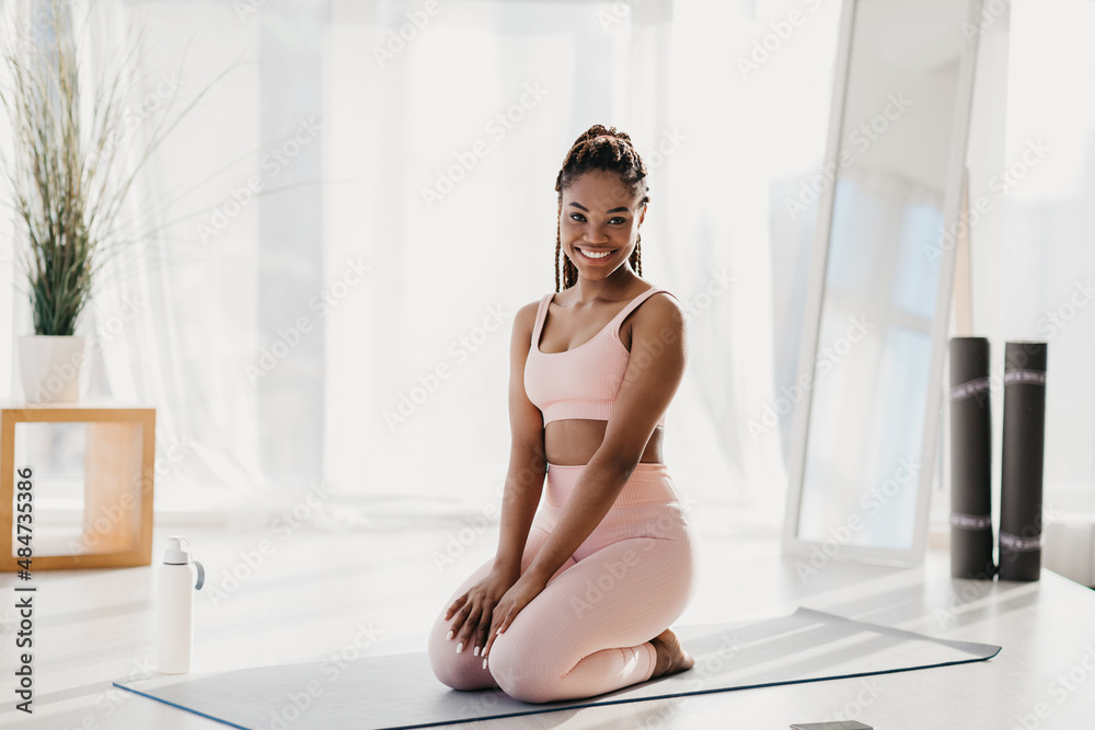 Smiling young black woman exercising at home, taking break, resting on yoga mat indoors, full length