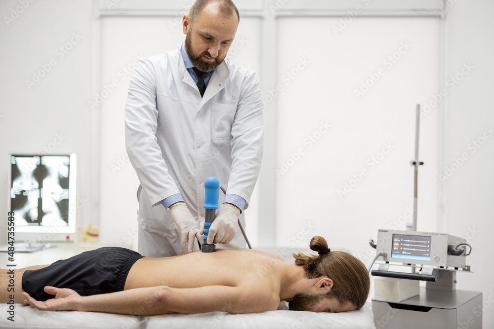 Senior doctor applies shock wave therapy with special medical equipment on men's back at medical office. Concept of non-invasive technology for treating pain in the musculoskeletal system
