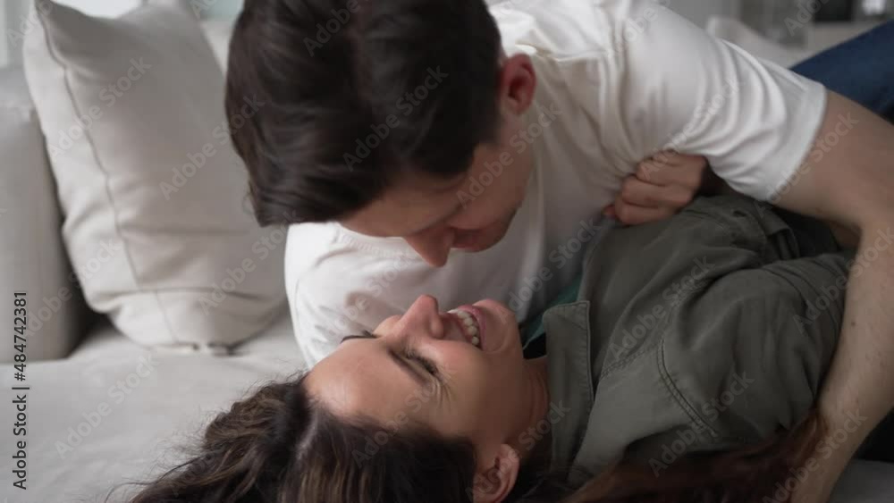 Happy newlyweds with dark hair have fun tickling and hugging lying on sofa and kissing softly with love in living room extreme closeup Happy newlyweds with dark hair have fun tickling and hugging lying on sofa and kissing softly with love in living room extreme closeup