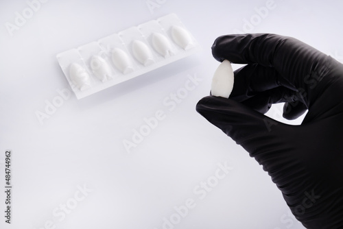 A hand in a black medical glove holds an anal suppository against the background of packaged anal suppositories. The concept of treating anal disease, hemorrhoids photo