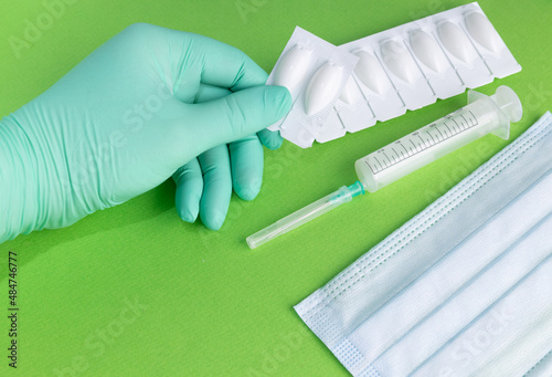 The hand holds vaginal suppositories on a green background. A hand in a medical glove holds anal suppositories, a medical mask and a syringe lie nearby. Concept photo