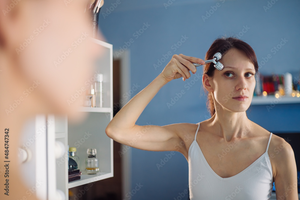 Young woman makes herself a face massage with massage face roller in front of mirror at home. Facial skin care lifting, anti age and beauty concept Young woman makes herself a face massage with massage face roller in front of mirror at home. Facial skin care lifting, anti age and beauty concept