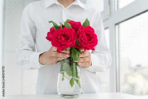 boy in white shirt puts red roses in big transparent vase, grreting and love concept, valentines day, photo