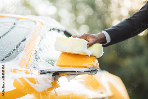 Closeup cropped image of hand of young African businessman with yellow sponge washing rearview mirror of his car at a self-serve car wash outdoors. Luxury car covered by foam.