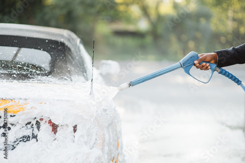 Car wash outdoors concept. Yellow car in foam. Car getting a wash with soap. Hand of African man with high pressure jet spraying the soap on yellow car at service station.