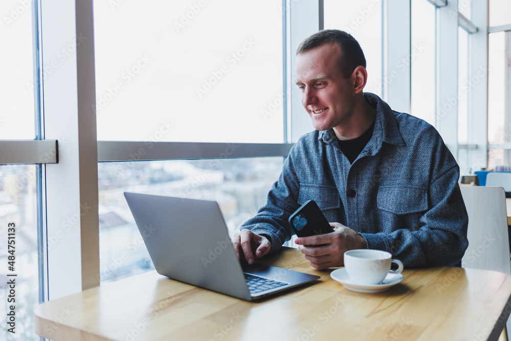 Successful happy business man is sitting at a table in a cafe, holding a cup of coffee and using a laptop. Successful happy business man is sitting at a table in a cafe, holding a cup of coffee and using a laptop.