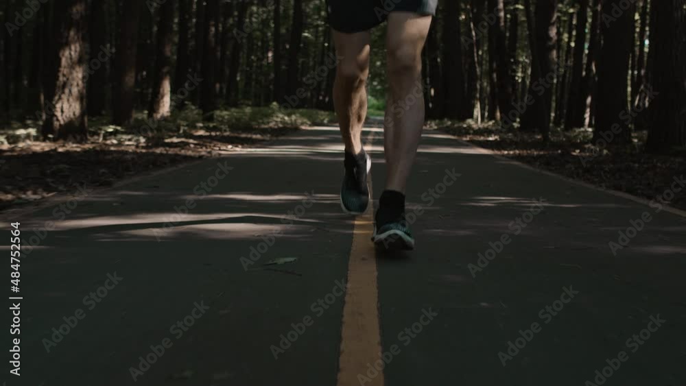 Athletic fit man with defined muscle legs on early morning training. Young man on workout plan preparation for marathon or competition run in park on asphalt road, track running Athletic fit man with defined muscle legs on early morning training. Young man on workout plan preparation for marathon or competition run in park on asphalt road, track running
