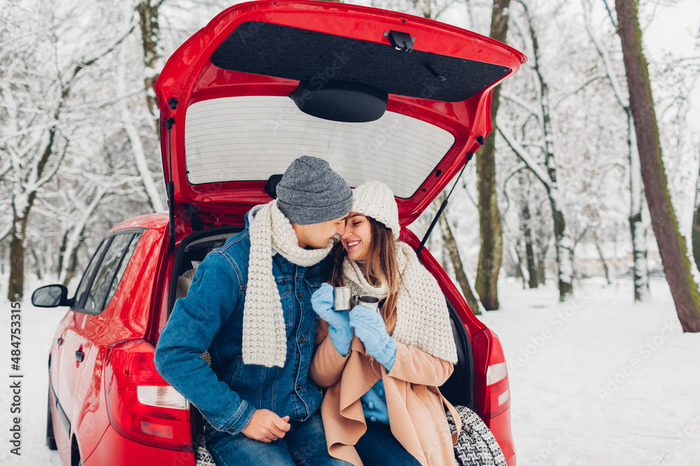 Valentine's day. Couple in love sitting in car trunk drinking hot tea in snowy winter forest. People hugging outdoors
