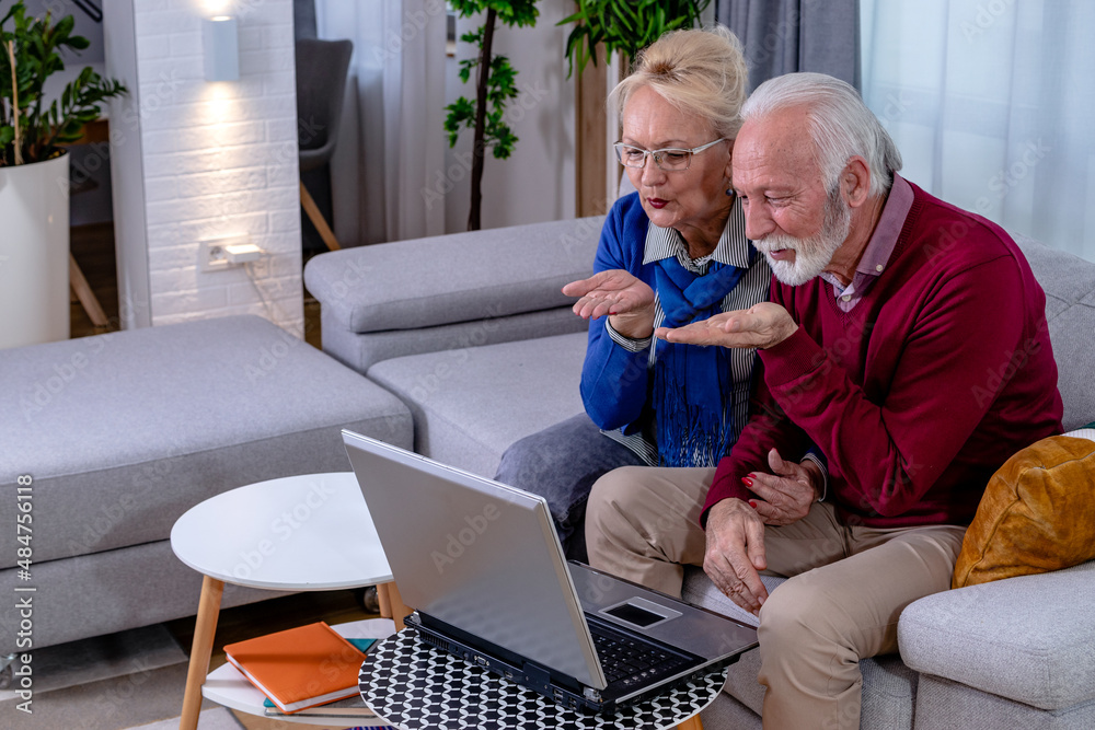 Lovely senior couple is sitting on a sofa and talking to their children and grandchildren via video call on a laptop in their living room. The elderly husband and wife are sending kisses at a camera.