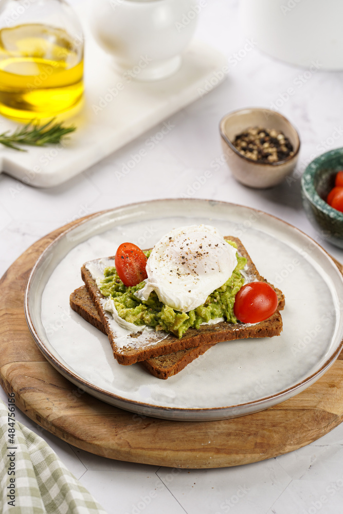Trendy lifestyle sandwich: protein bread slice with cream cheese, mashed avocado, cherry tomatoes and poached egg on white scandi plate, light setting