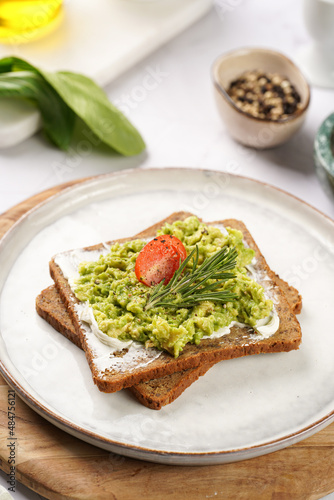 Trendy lifestyle sandwich: protein bread slice with cream cheese, mashed avocado, cherry tomatoes and rosemary on white scandi plate, light setting