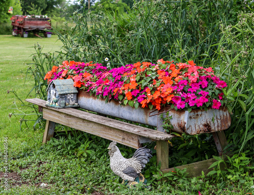 An old chicken feeder filled with colorful impatiens and decorative rural ornaments and an Amish farm wagon sits in the background.  photo