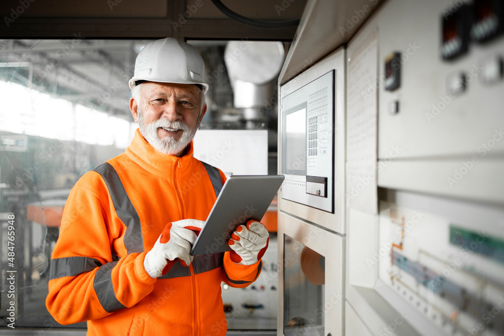 Portrait of professional factory worker or engineer in high visibility jacket and hardhat standing in control room of industrial production hall. Factory interior. Portrait of professional factory worker or engineer in high visibility jacket and hardhat standing in control room of industrial production hall. Factory interior.