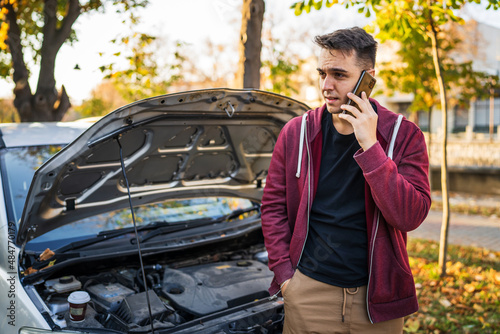 One young adult caucasian man standing by his vehicle with open hood and broken failed engine holding a phone calling towing service for help on the road Roadside assistance concept in autumn day