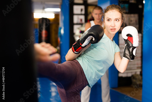 Blonde woman kicking punchbag during group kickboxing training. Her trainer standing behind and observing.