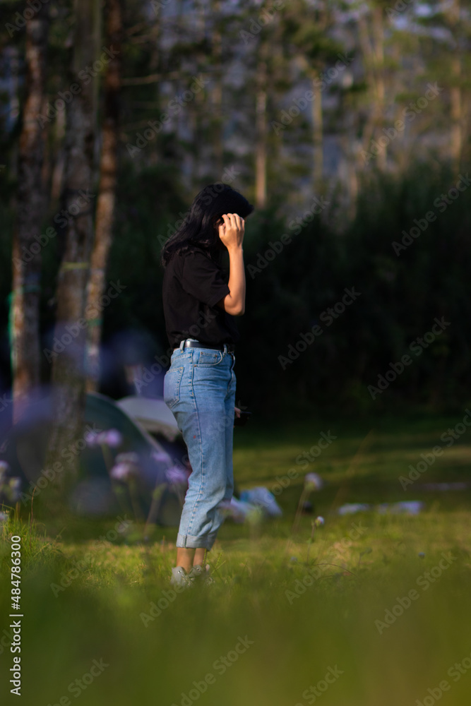 A relaxing girl with blue jeans is standing on green grass with blooming flowers while touching her hair
