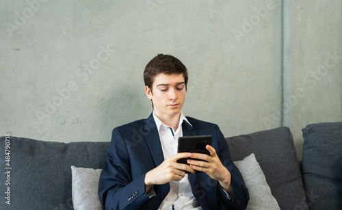 caucasian businessman working with tablet, wear suit, sitting on sofa, copy space at grey wall background. Young man work business with home office background and copy space