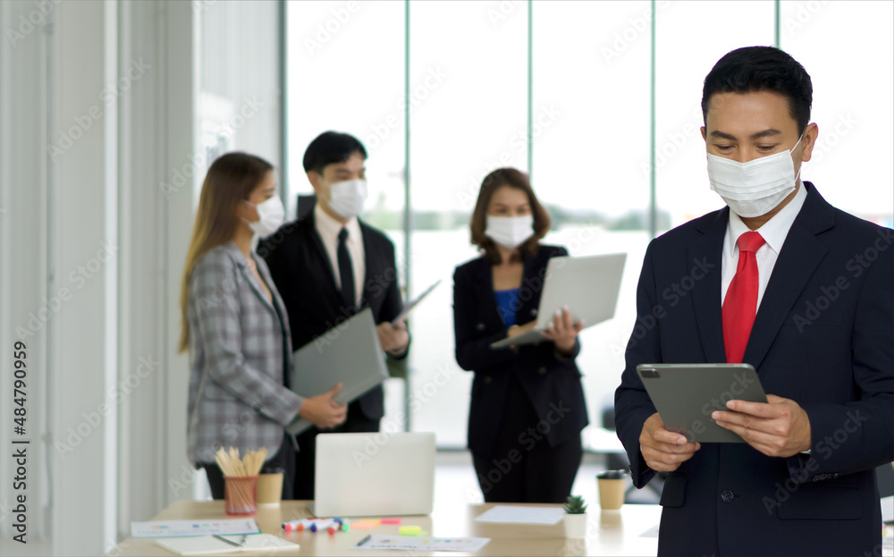 Asian businessman in black suit standing with tablet computer in his hand. The team is preparing for the meeting in the background. Everyone wears a mask. Concept of leadership. Asian businessman in black suit standing with tablet computer in his hand. The team is preparing for the meeting in the background. Everyone wears a mask. Concept of leadership.