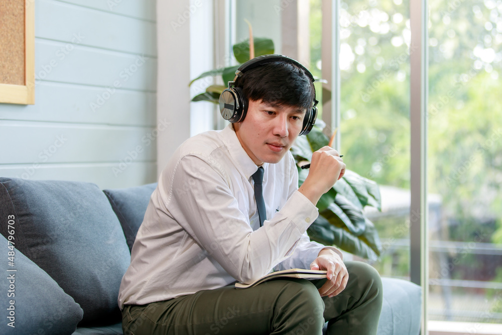 Asian young handsome professional successful male businessman employee in formal business shirt and necktie sitting on cozy sofa listening to streaming music online browsing internet .
