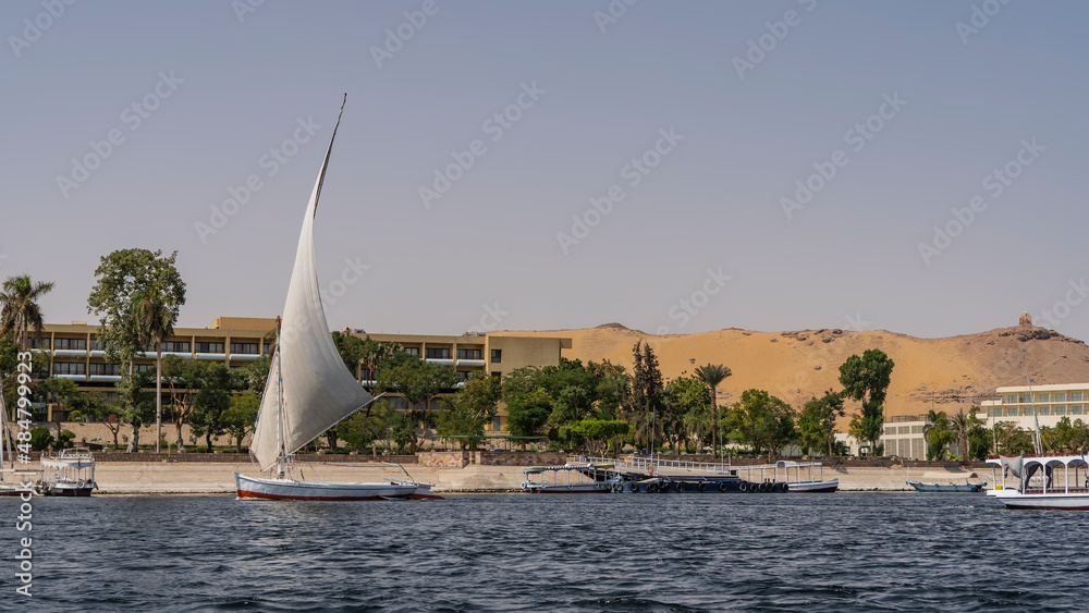 There are tourist boats and a felucca with a raised sail on the shore of the Nile. Coastal green vegetation. A sand dune against a blue sky. Egypt. Aswan