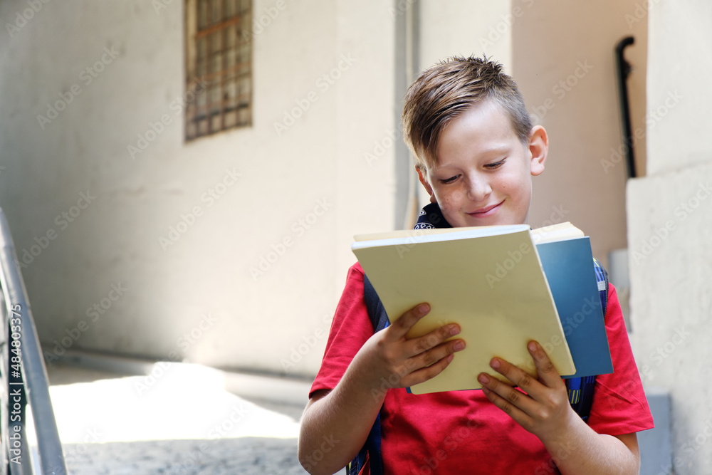 Young boy, elementary school student, reading books after school on his way home, enjoying a sunny day with his favorite literature Young boy, elementary school student, reading books after school on his way home, enjoying a sunny day with his favorite literature