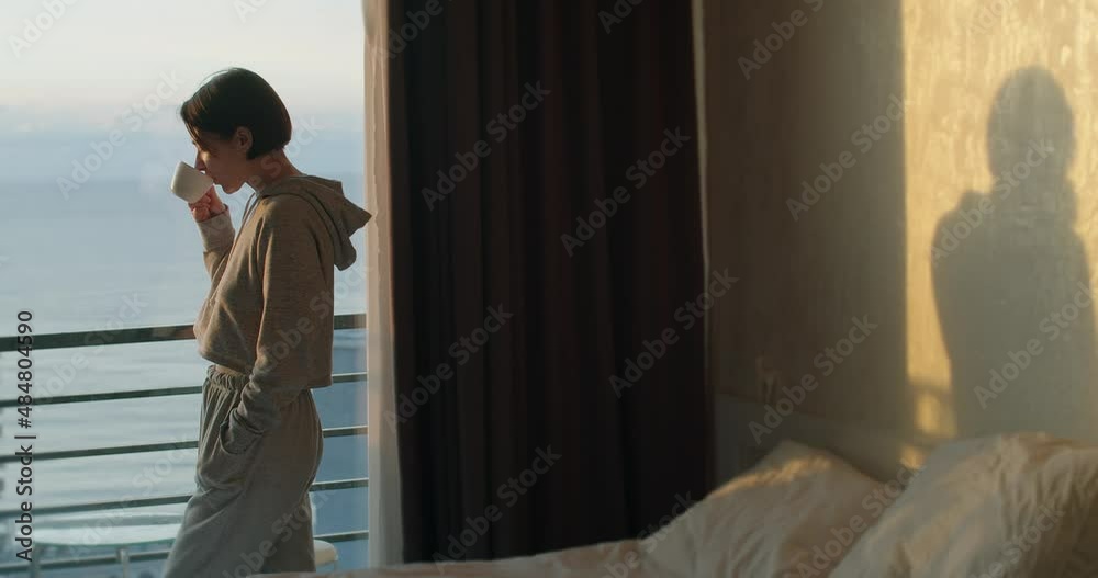 Attractive woman standing by window with cup of tea and enjoying beautiful view. Young girl drinking coffee watching sunset and relaxing. Peaceful evening at home Attractive woman standing by window with cup of tea and enjoying beautiful view. Young girl drinking coffee watching sunset and relaxing. Peaceful evening at home
