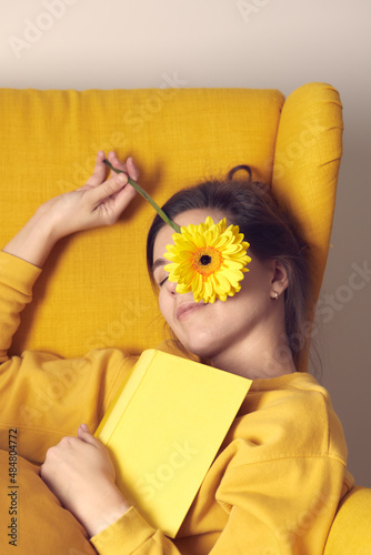 A beautiful girl dreams of sitting in a yellow chair with a book and a flower in her hands. Front view.