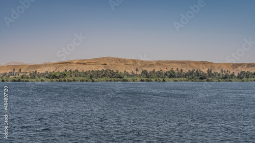 Along the bank of the Nile, thickets of lush green grass and palm trees are visible. A high sand dune against a clear azure sky. Ripples on the blue calm water. Egypt