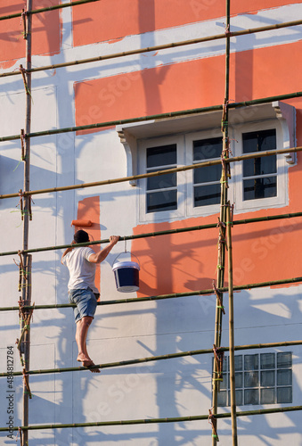 Low angle view of Asian builder worker on wooden scaffolding is painting wall outside of the old house in vertical frame
