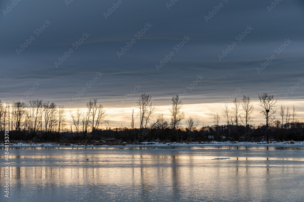 sunrise by the river on a winter morning with burning cloud behind the silhouette of trees on the shore over the horizon