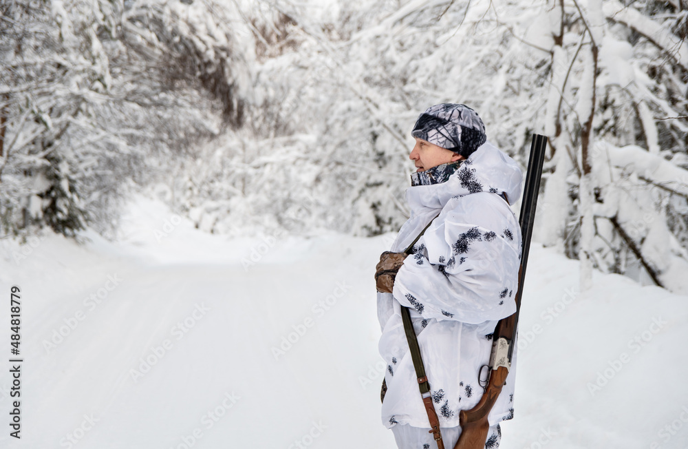 A male hunter in a white comfly suit in a winter forest with a gun on his shoulder. Copy space.