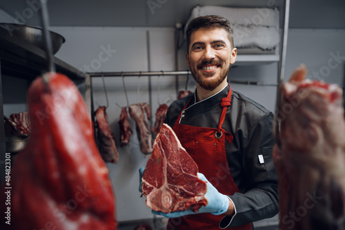 Young butcher holding raw meat steaks in fridge of grocery shop