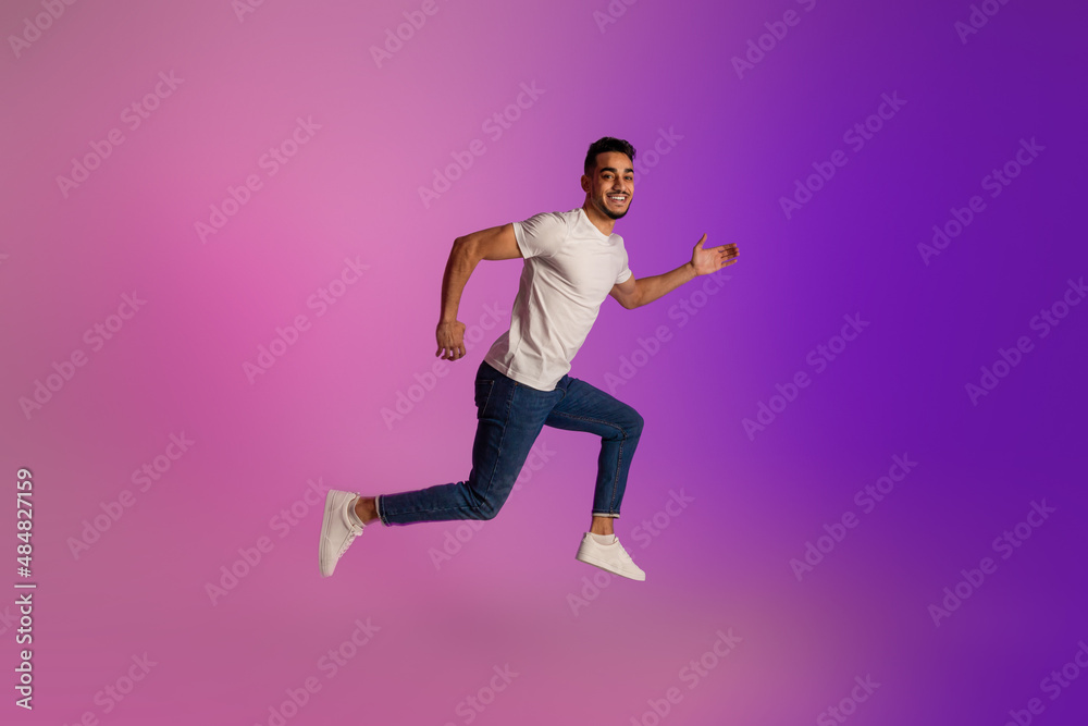 Full length portrait of positive young Arab man in casual wear jumping in neon light, copy space
