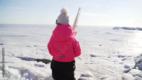 litttle girl skier in pink jacket with white skis on her shoulder goes to the frozen sea and snow photo