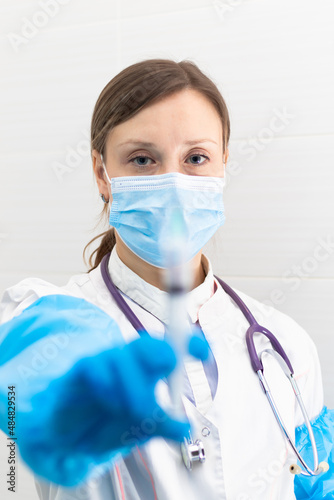 Young woman doctor in a medical mask and gloves with a syringe in her hand and a stethoscope in a hospital during a coronavirus pandemic. Selective focus. Portrait