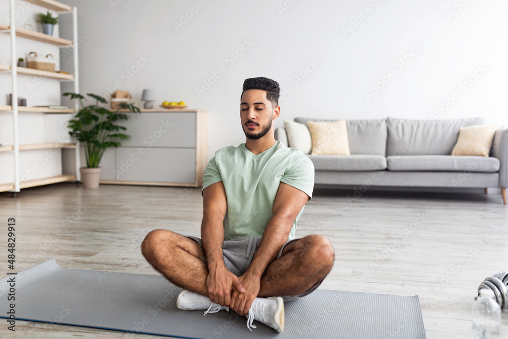 Peaceful millennial Arab guy sitting in lotus pose, meditating with closed eyes on mat at home, copy space