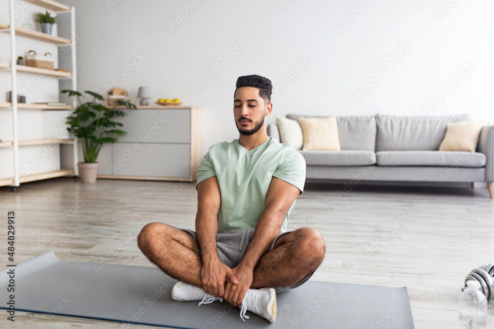 Fototapeta premium Peaceful millennial Arab guy sitting in lotus pose, meditating with closed eyes on mat at home, copy space