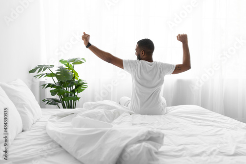 Calm millennial african american guy sitting on bed stretching body and enjoying good morning
