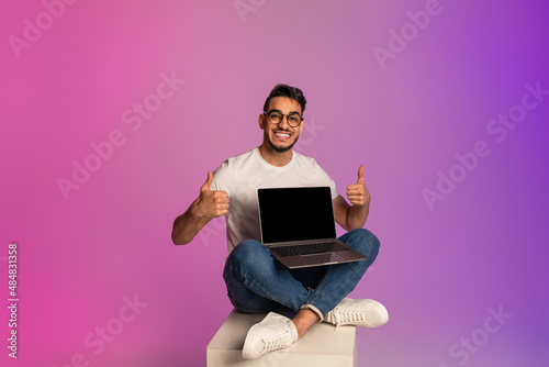 Young Arab guy holding laptop with empty screen, showing thumb up, recommending new website in neon light. Mockup