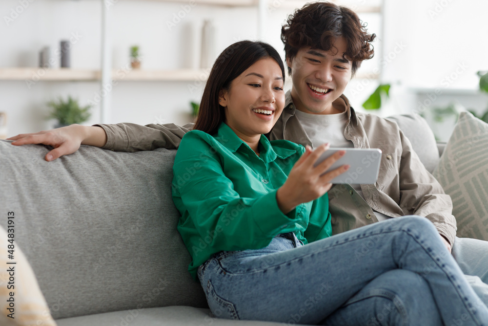 Obraz premium Millennial Asian couple using smartphone, relaxing on weekend, sitting on couch together at home, copy space