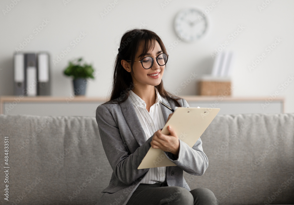 Focused arab female psychologist sitting on couch and writing in clipboard, offering professional counseling services Focused arab female psychologist sitting on couch and writing in clipboard, offering professional counseling services