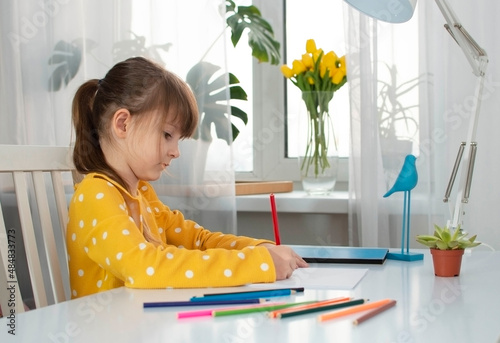 A girl in a yellow dress draws in her room at her desk with colored pencils. The concept of children's creativity and development. Selective focus
