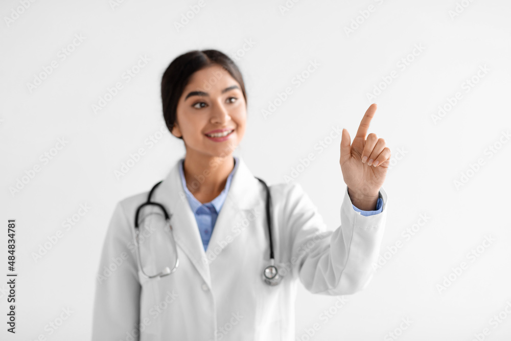 Smiling pretty young hindu woman doctor in white coat with stethoscope points finger to blank space Smiling pretty young hindu woman doctor in white coat with stethoscope points finger to blank space