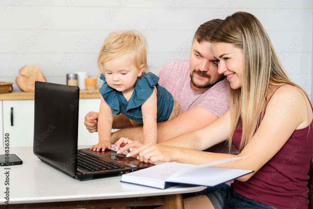Young parents and a small child sit at a table, smile and look at a laptop. High quality photo Young parents and a small child sit at a table, smile and look at a laptop. High quality photo