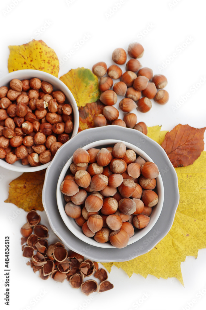 Whole hazenuts in a bowl with cracked hazenuts and autumnal leaves on white background Whole hazenuts in a bowl with cracked hazenuts and autumnal leaves on white background