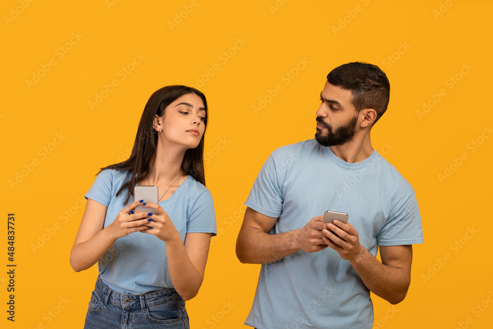 Fototapeta premium Privacy concept. Arab lady trying to look at her boyfriend's smartphone, guy hiding gadget, yellow studio background